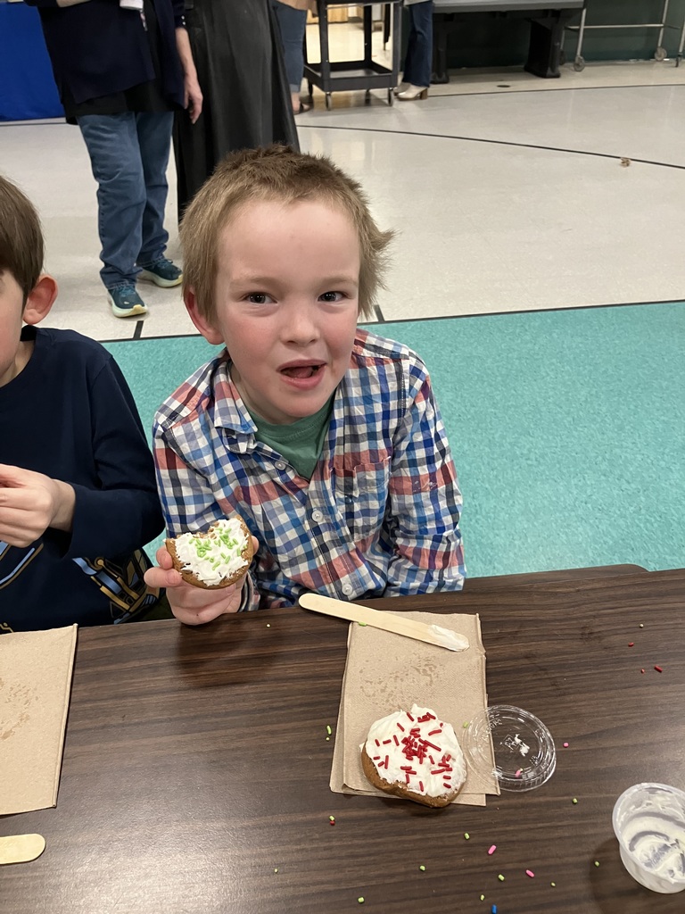 A boy in a blue and white plaid shirt sits at a dark wood table, holding a decorated cookie with green sprinkles that has a large bite taken out of it. Another cookie with red sprinkles sits on a napkin in front of him. He is looking at the camera with a cheerful, mid-snack expression.
