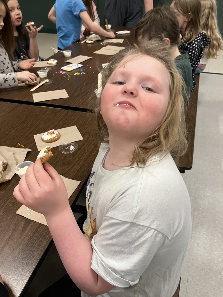 A young girl with blonde hair tilts her head back and smiles while holding a piece of a decorated cookie. She has a bit of white frosting on her lip. Behind her, other students are sitting at the long tables, busy decorating or eating their own cookies. 
