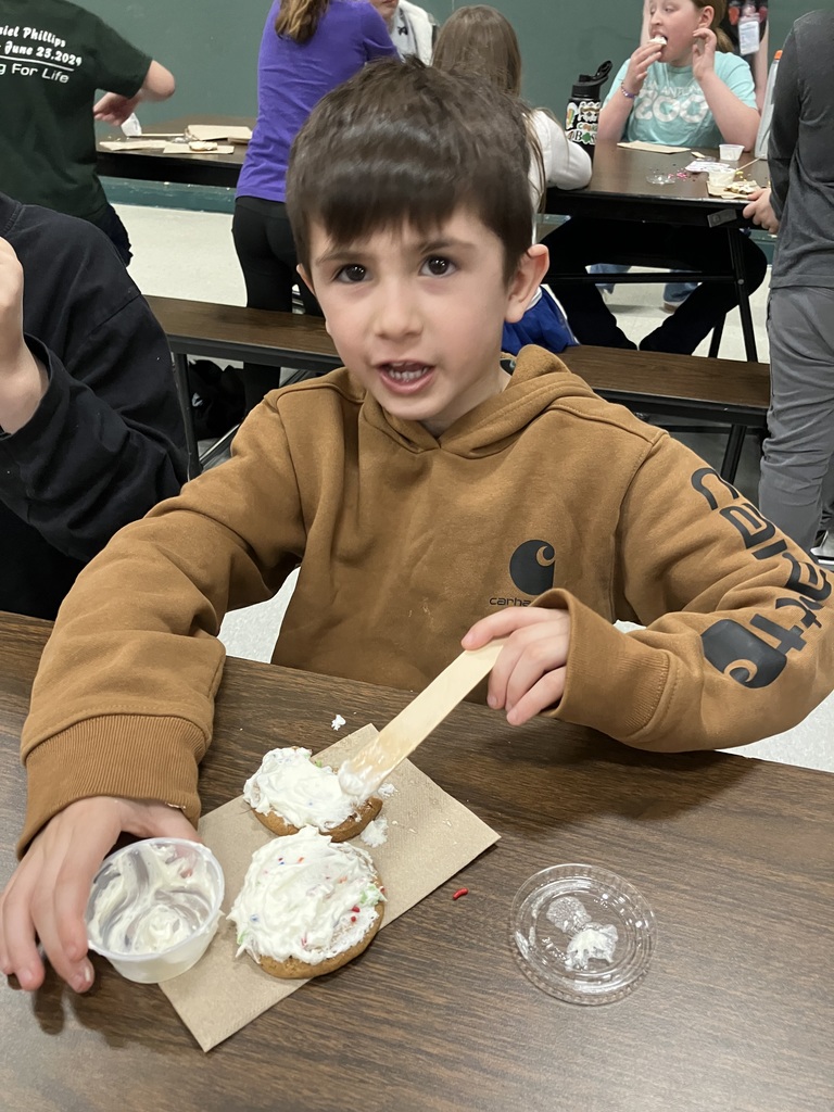  A boy in a brown Carhartt hoodie uses a wooden stick to spread thick white frosting onto two cookies sitting on a brown paper napkin. He is looking toward the camera with an open-mouthed, excited expression.  