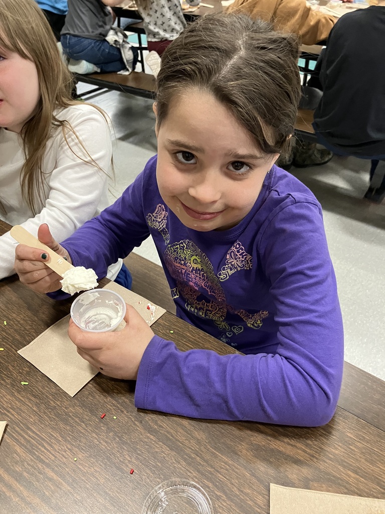  A young girl with dark hair and a purple long-sleeved shirt smiles at the camera. She is sitting at a cafeteria table and holding a wooden craft stick covered in a large dollop of white frosting, which she has just scooped from a small plastic cup.  