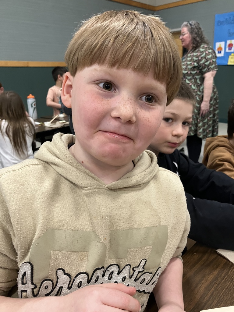 pg A close-up of a young boy with light brown hair and freckles wearing a tan "Aeropostale" hoodie. He has a playful, tight-lipped expression as if he's just taken a bite of a treat. Other children and an adult in a patterned dress are visible in the background of the gymnasium.  