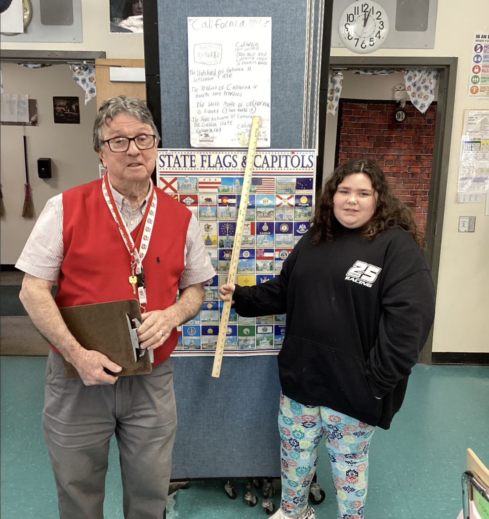 Teacher holds a clipboard while standing next to a student holding a yardstick, with a California project poster displayed above.