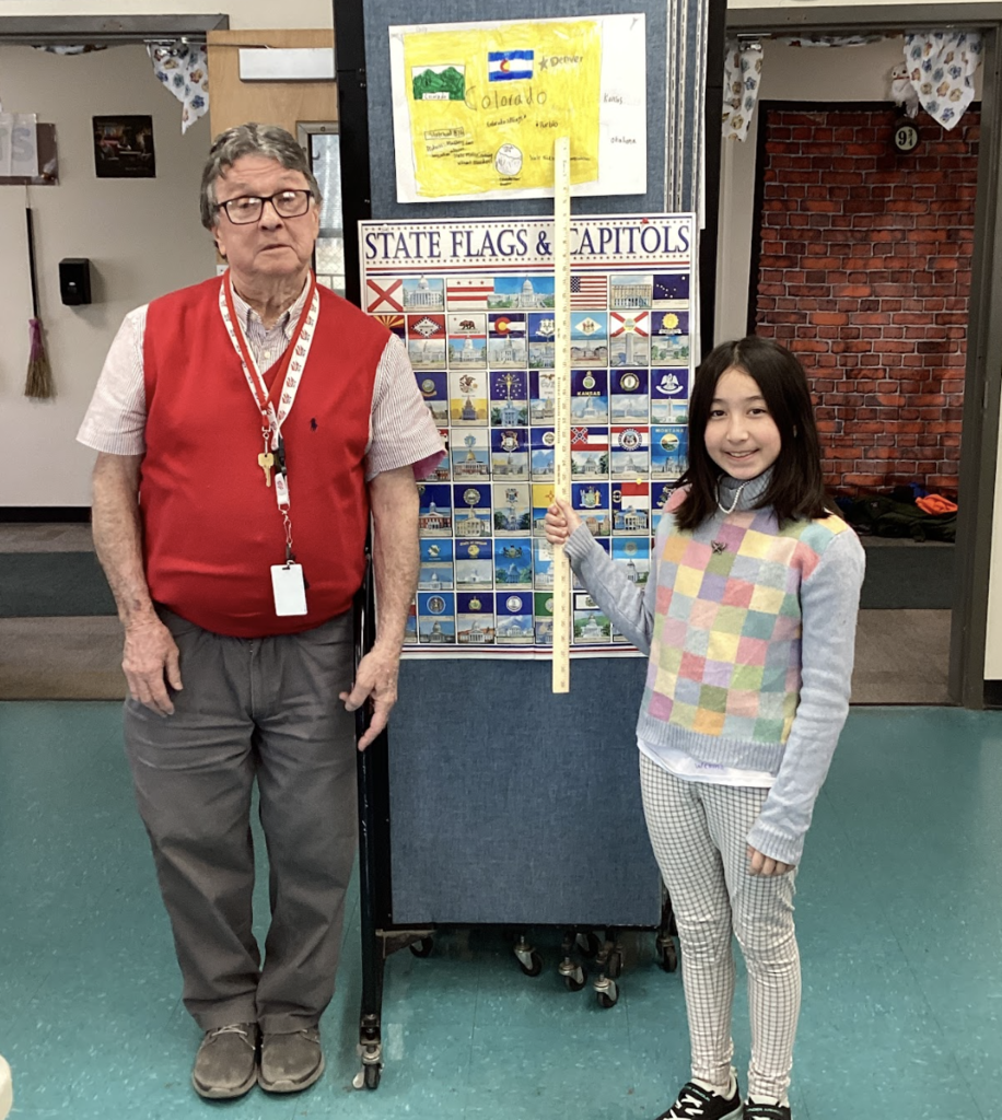Teacher stands with a student holding a yardstick in front of a state display board, with a Colorado project poster above.