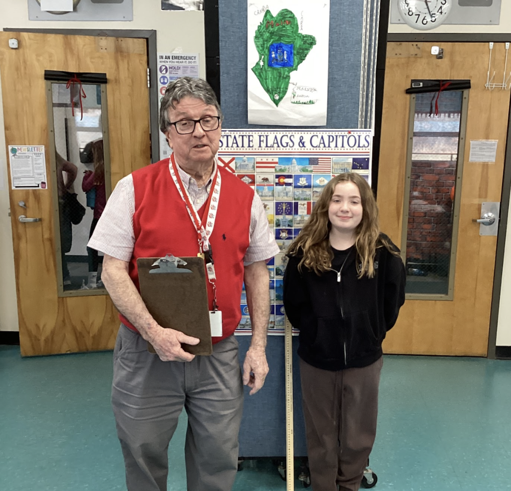 Older male teacher holding a clipboard stands beside a student in front of a “State Flags & Capitals” display, with a student-made state project poster mounted above.