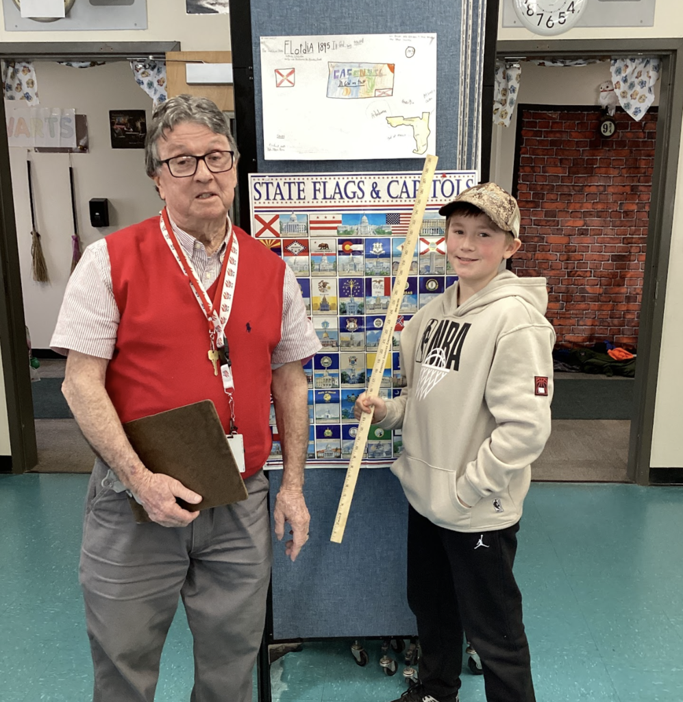 Teacher and student stand in front of a state display board, with a Florida project poster above and a yardstick visible.