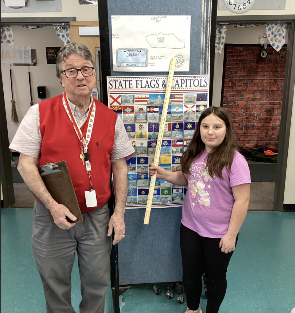 Teacher and student stand beside a state display board, with a Delaware project poster above and a yardstick held upright.