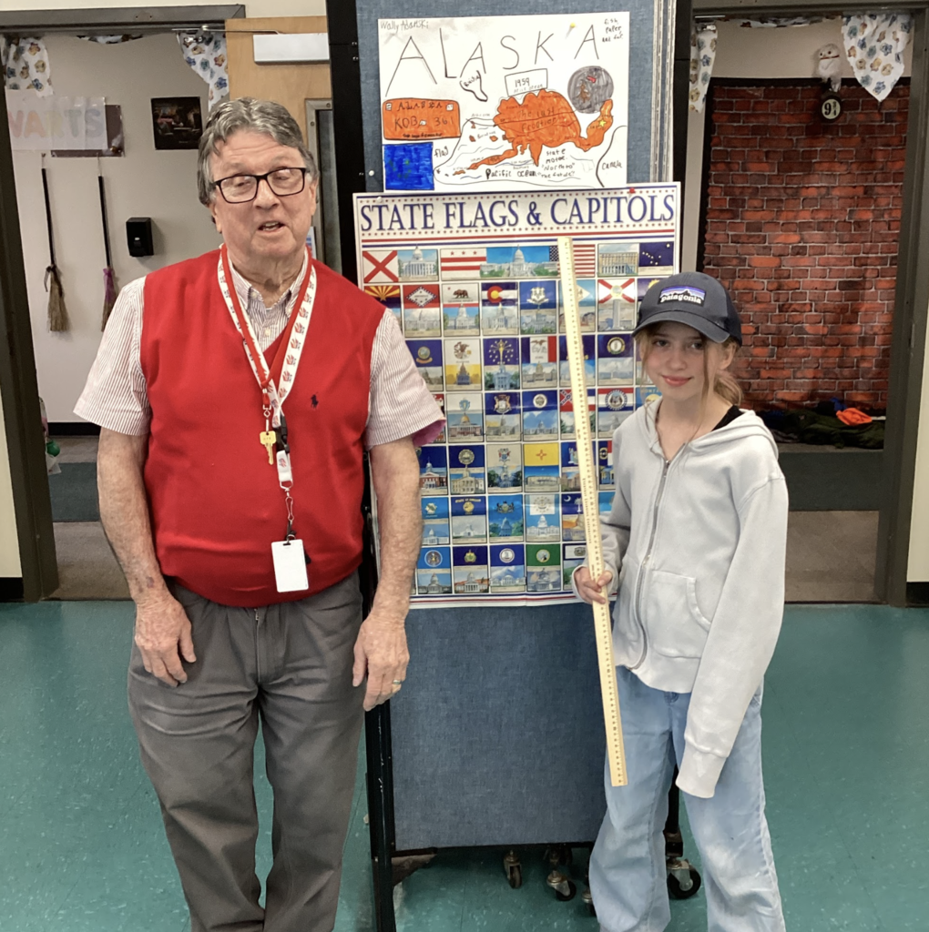 Older male teacher in a red vest stands beside a student holding a yardstick in front of a “State Flags & Capitals” display and an Alaska project poster.