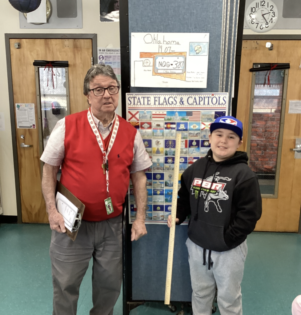 Teacher stands beside a student holding a yardstick in front of a state display board, with an Oklahoma project poster above.