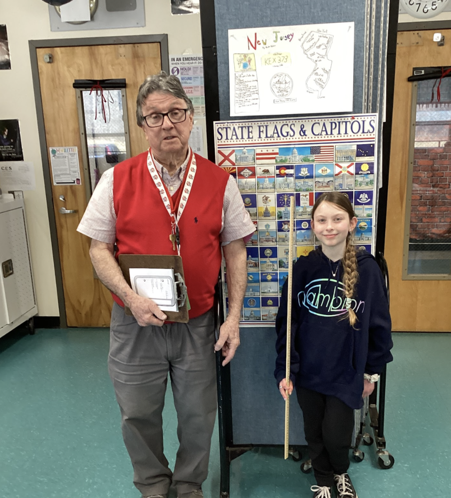 Teacher stands with a student holding a yardstick in front of a state display board, with a New Jersey project poster above.
