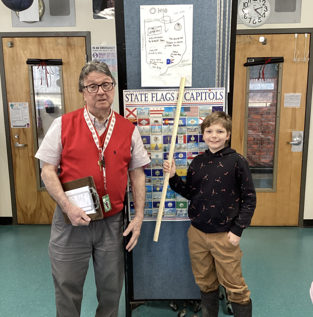 Teacher and student stand in front of a state display board, with an Ohio project poster above and a yardstick visible.