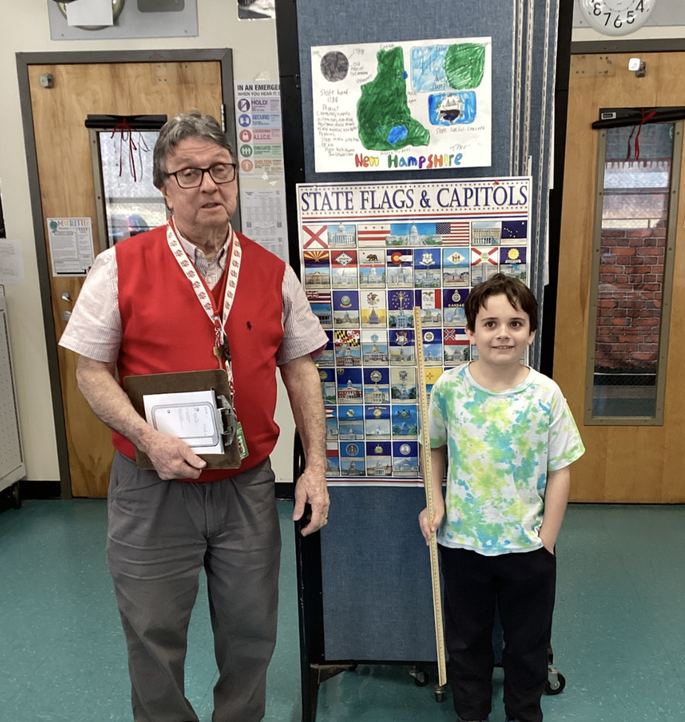 Teacher and student stand beside a state display board, with a New Hampshire project poster above and a yardstick held upright.