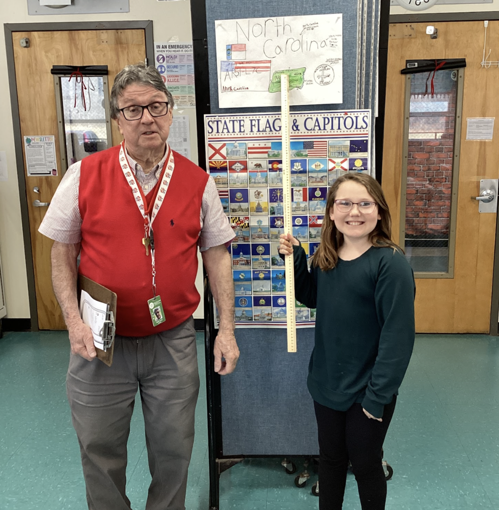 Teacher stands next to a student holding a yardstick in front of a state display board, with a North Carolina project poster above.
