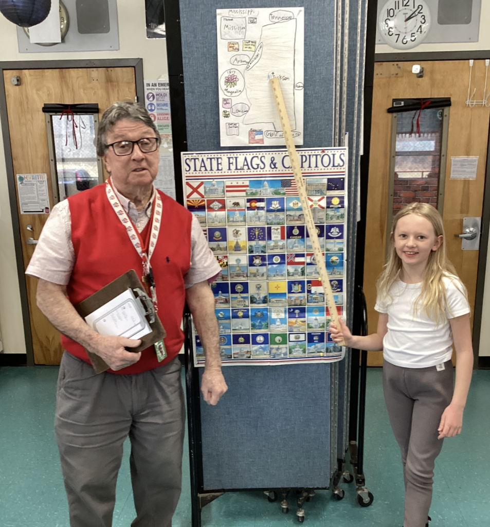 Teacher stands next to a student holding a yardstick in front of a state display board, with a Mississippi project poster above.
