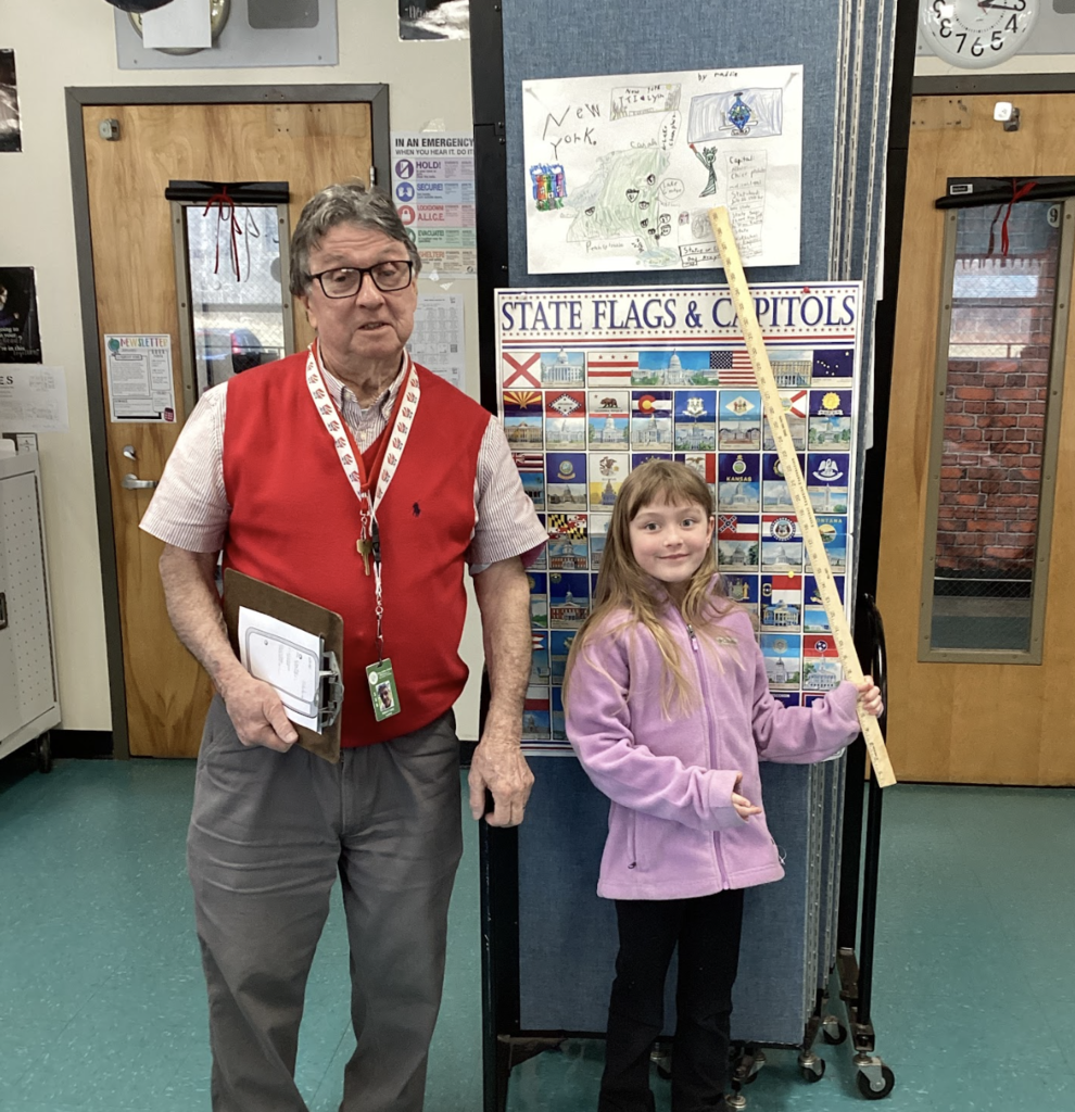 Teacher and student stand beside a state display board, with a New York project poster above and a yardstick held upright.