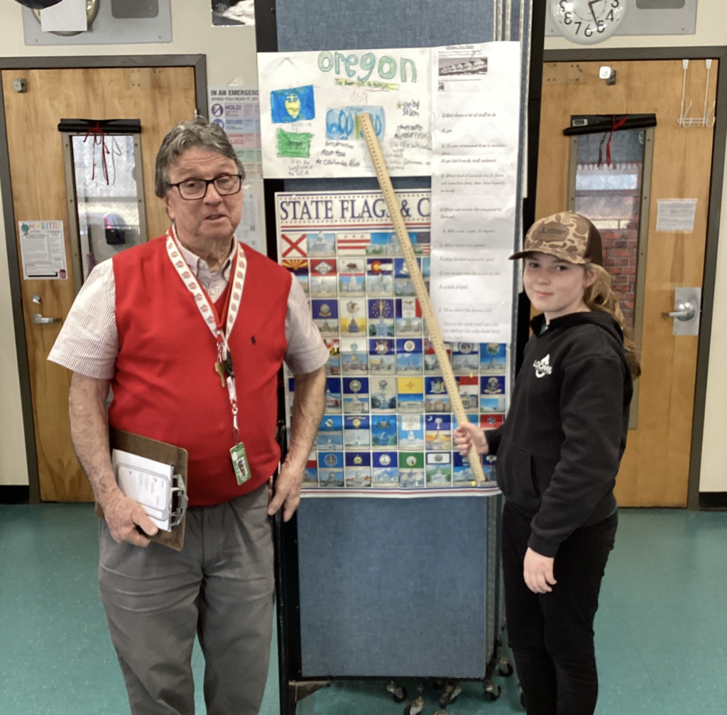 Teacher and student stand in front of a state display board, with an Oregon project poster above and a yardstick pointing upward.
