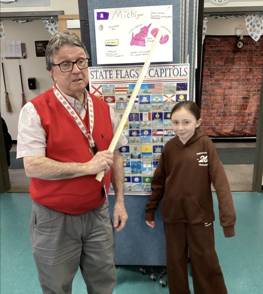 Teacher holds a yardstick beside a student in front of a “State Flags & Capitals” display, with a Michigan project poster above.