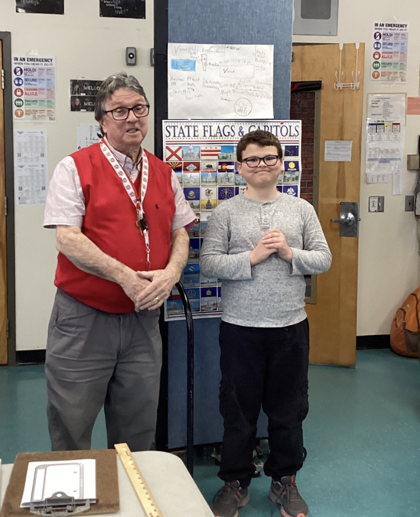 Teacher and student stand near a table with a yardstick, in front of a state display board.