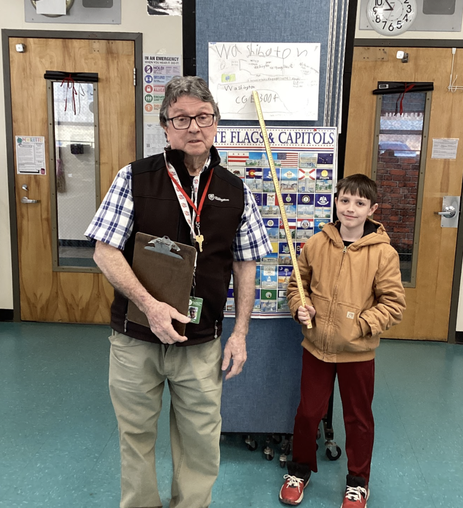 Teacher and student stand with a yardstick in front of a Washington project and state display board.