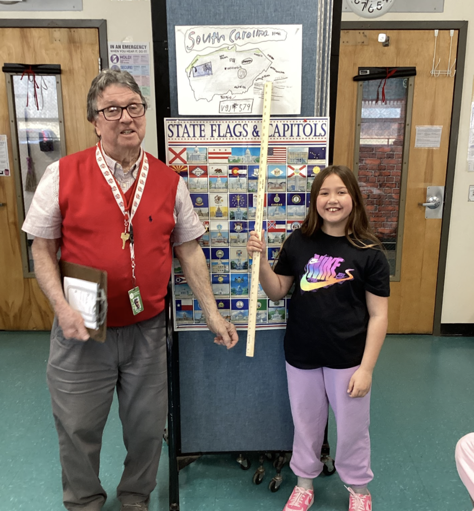 Teacher and student hold a yardstick beside a “State Flags & Capitals” board with a South Carolina project above.