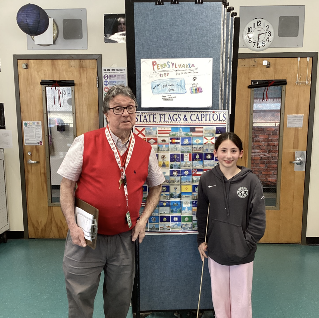 Teacher and student stand by a “State Flags & Capitals” board with a Pennsylvania project displayed above.