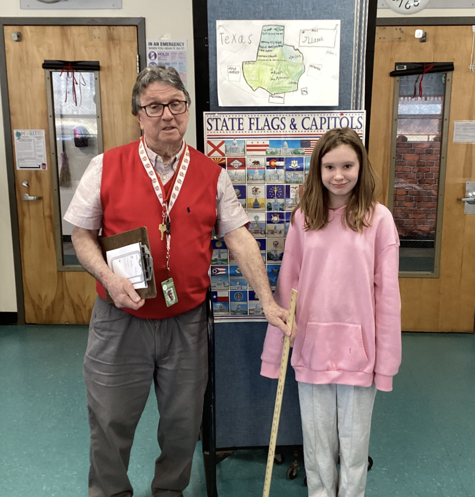 Teacher and student stand with a yardstick in front of a Texas project and state display board.