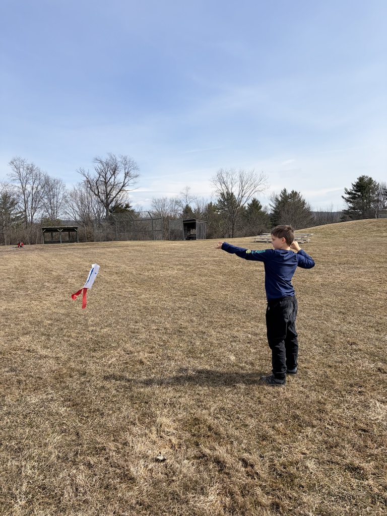 A young boy in a long-sleeved navy shirt and black pants stands in a dry, grassy field on a clear day. He holds a thin string connected to a small, homemade white kite with a red streamer tail, which is flying several feet above the ground to his left.