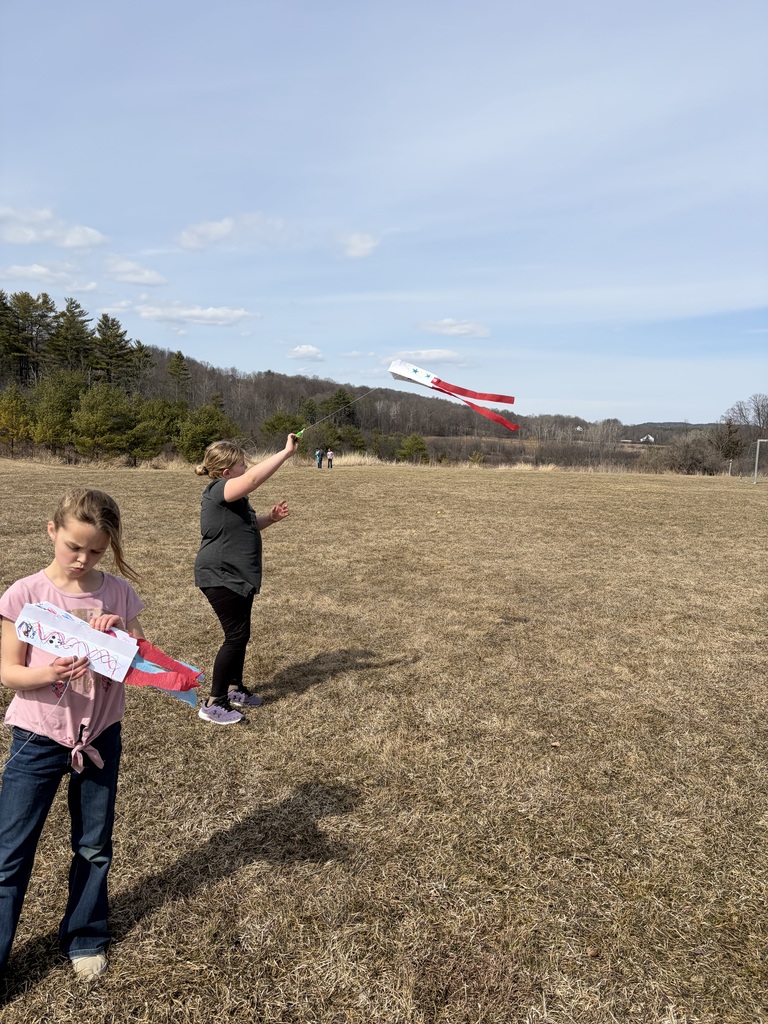 A photograph of two young girls in a dry, grassy field with a line of bare trees in the distance. The girl on the right is looking up and flying a small kite with two long red tails on a string, which is catching the wind. The girl on the left is standing holding her own folded-up red and blue paper kite. Both girls have their shadows cast on the grass. The sky is bright blue with a few light, scattered clouds.