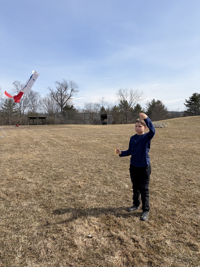 A young boy in a long-sleeved navy shirt and black pants stands in a dry, grassy field on a clear day. He holds a thin string connected to a small, homemade white kite with a red streamer tail, which is flying several feet above the ground to his left. 