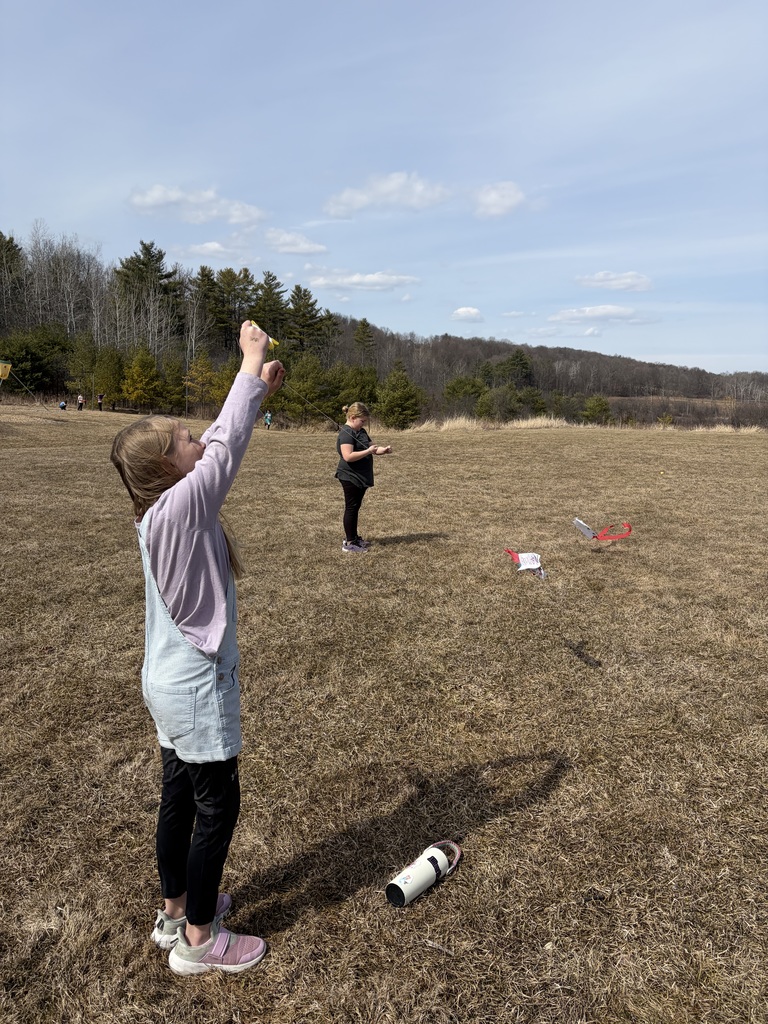 A young boy in a long-sleeved navy shirt and black pants stands in a dry, grassy field on a clear day. He holds a thin string connected to a small, homemade white kite with a red streamer tail, which is flying several feet above the ground to his left.