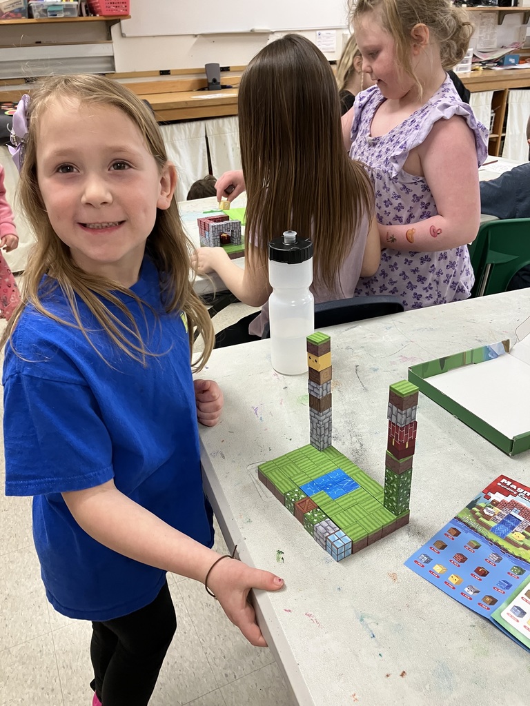 A young girl in a blue t-shirt smiles at the camera next to a tall structure she built using magnetic Minecraft-style blocks on a classroom table