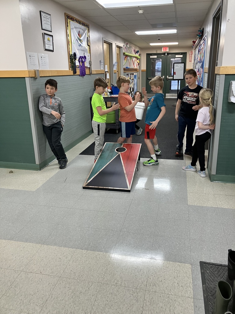  A group of elementary school children playing cornhole in a school hallway. Two boys are high-fiving over a wooden cornhole board painted with a teal and red geometric design. 