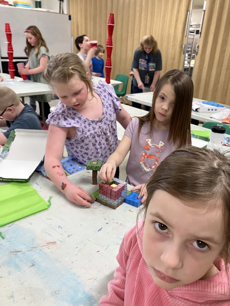 A young girl in a purple floral shirt works collaboratively with another student to build a small house out of magnetic blocks. A girl in a pink sweater is in the foreground.