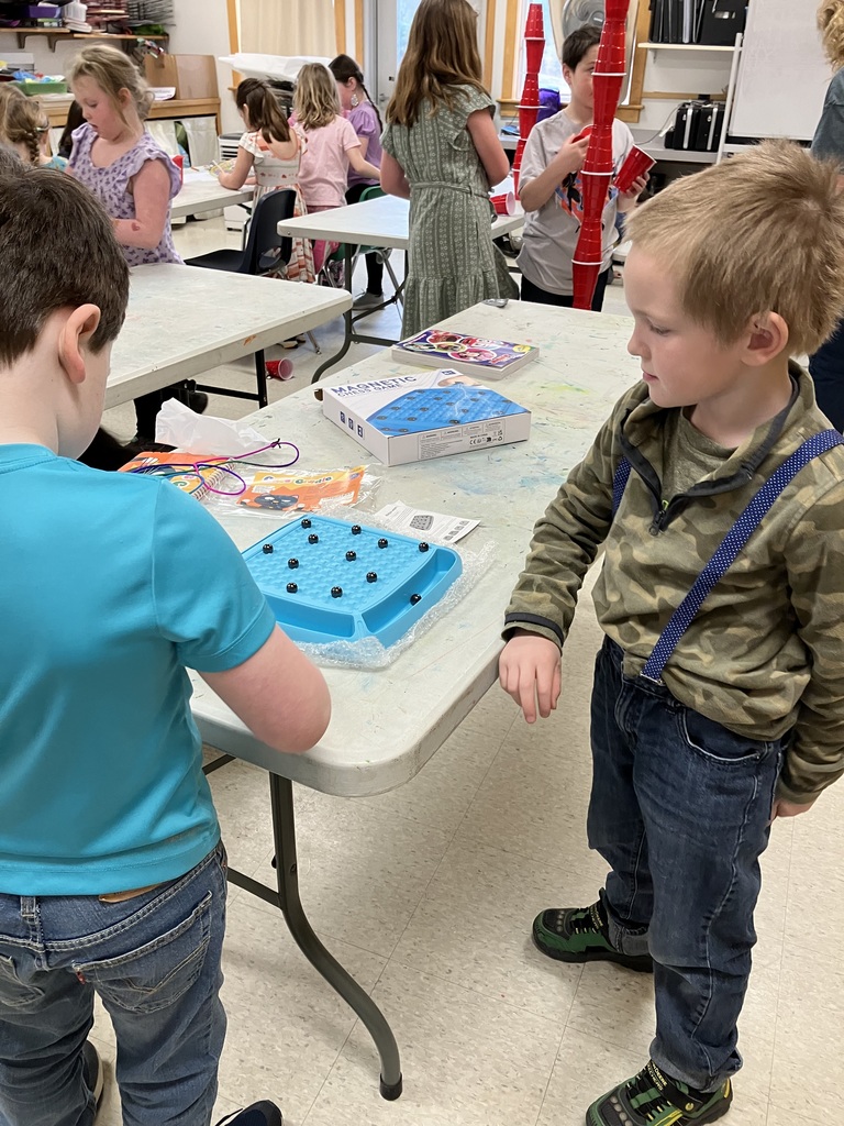 Two young boys standing at a classroom table, looking intently at a blue "Magnetic Chess Game" board. In the background, other children are working on various activities.  