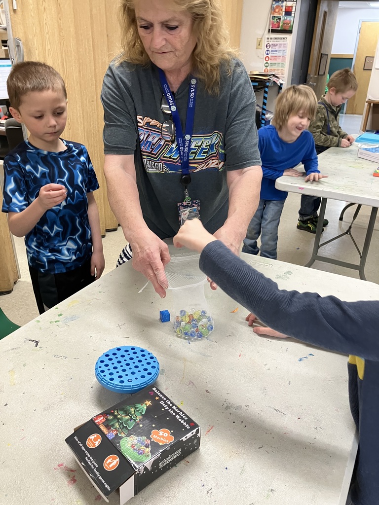 An adult supervisor assists a young boy with a marble game called "Balance the Marbles: Defy the Wobble." The teacher holds a mesh bag of colorful marbles over a blue perforated game base.