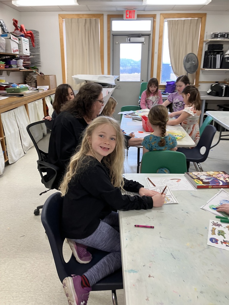 : A young girl with long blonde hair sits at a classroom table, smiling at the camera while coloring a drawing with a crayon. In the background, other students and a teacher work at separate tables.