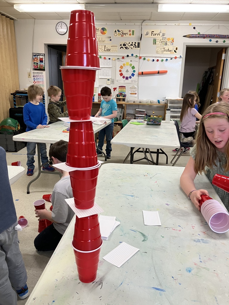  A close-up of a tall engineering project where red plastic cups are stacked vertically, separated by white index cards to create a multi-level tower on a classroom table.  IMG_3186.jpg: An adult supervisor assists a young boy with a marble game called "Balance the Marbles: Defy the Wobble." The teacher holds a mesh bag of colorful marbles over a blue perforated game base.