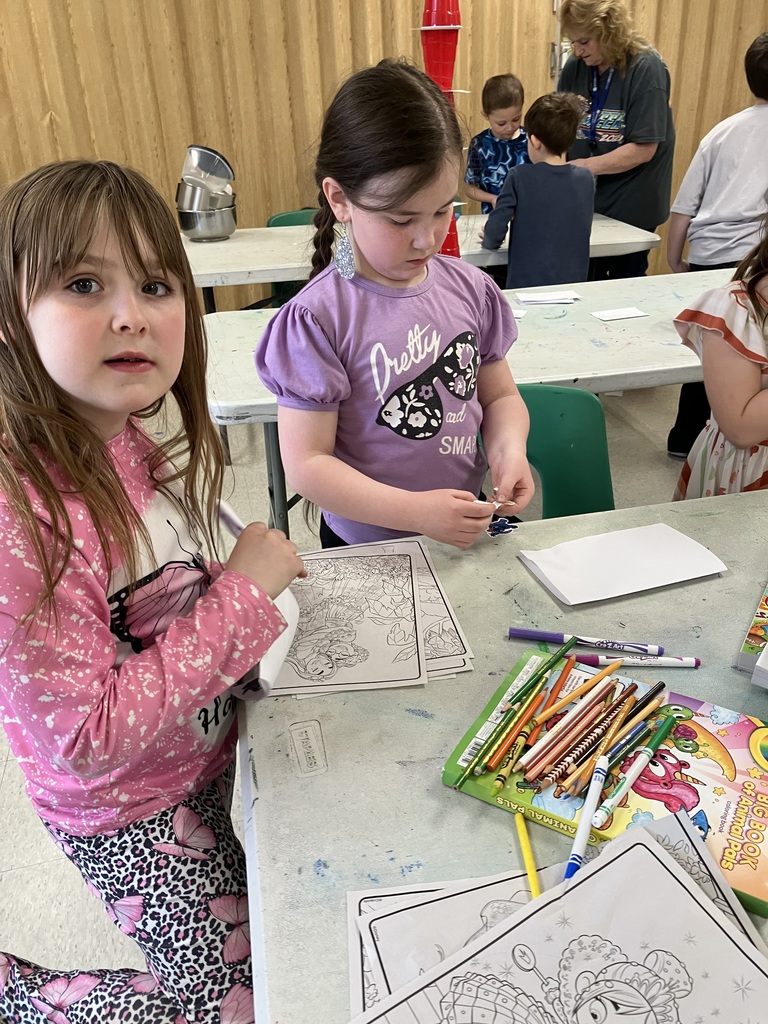 A girl in a pink butterfly-print shirt looks toward the camera while another girl in a purple "Pretty and Smart" t-shirt focuses on a sticker or craft project at a busy classroom table. 