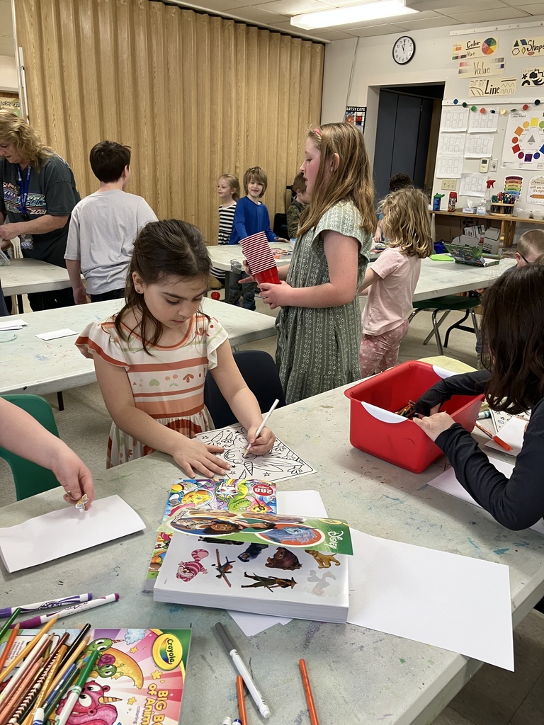 A wide shot of a classroom activity center. A girl in a striped dress is coloring, while another student nearby holds a stack of red plastic cups. Educational posters about color, shape, and line are visible on the wall. 