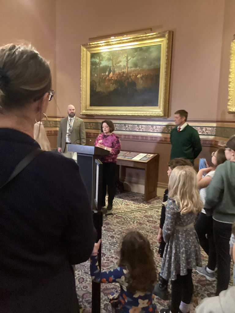 A speaker stands at a podium in a historic room while children and adults listen during a presentation.