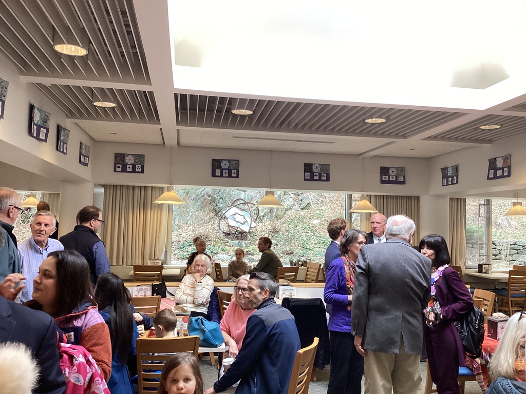 A large group of adults and children gather in a bright room, talking and viewing artwork displayed on the walls.