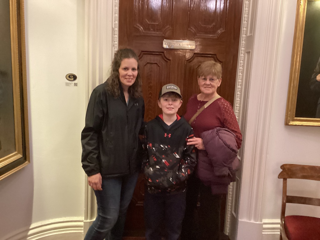 A student stands between two adults in a hallway inside the State House, posing in front of a wooden door.