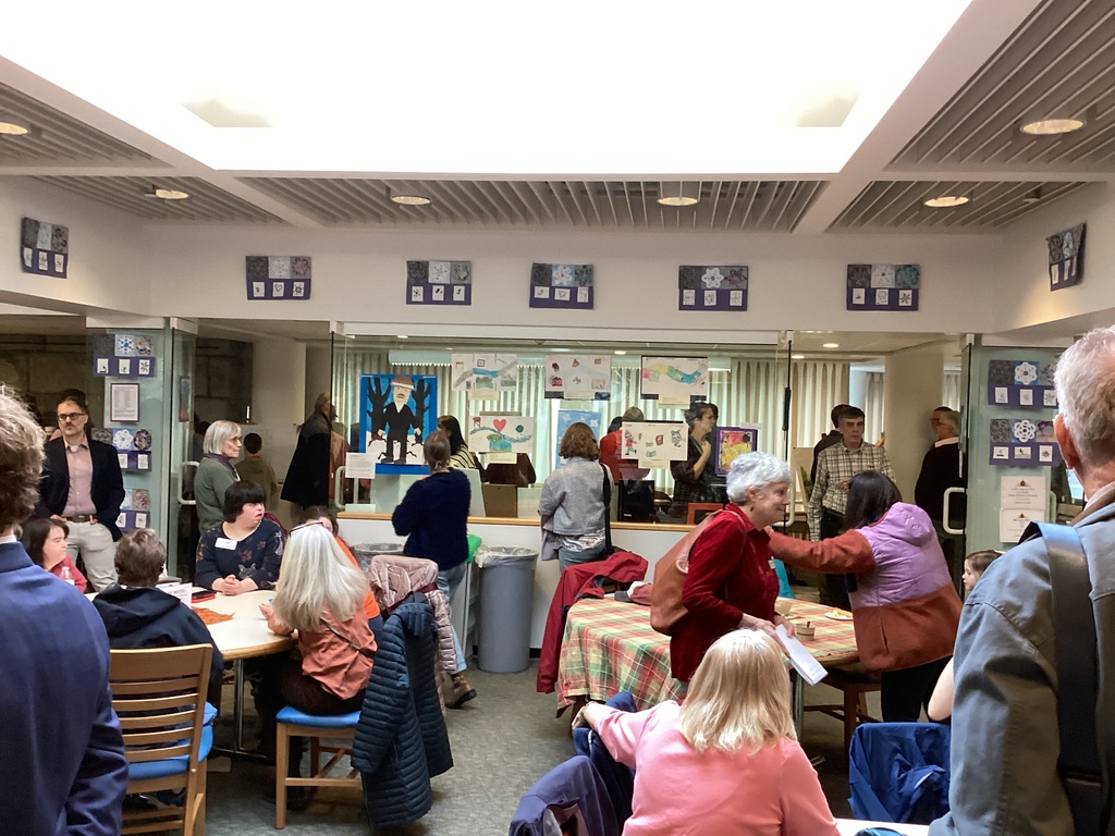 Families and community members sit and stand in a cafeteria-style space, looking at student artwork displayed behind glass.