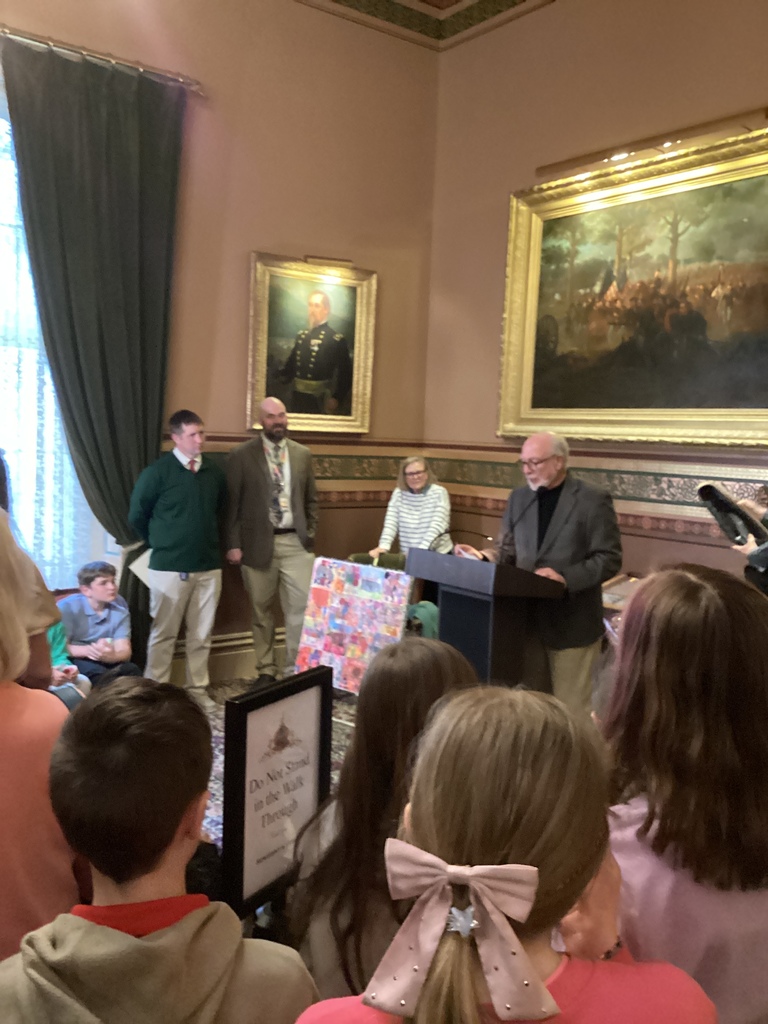 Another speaker addresses a group of students and adults in a historic room with framed paintings on the walls.