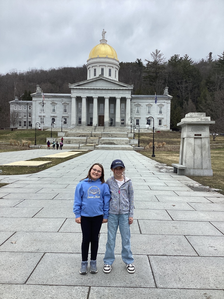 Two elementary-aged students stand smiling in front of the Vermont State House with its gold dome in the background.