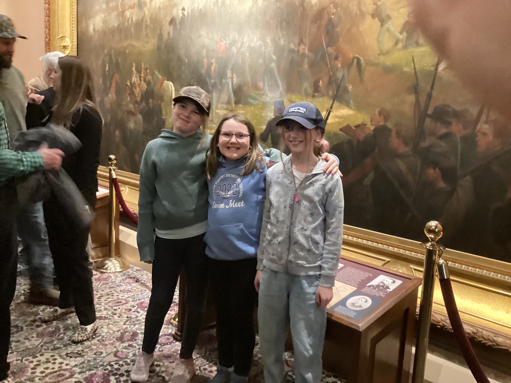 Three students smile and pose together in front of a large historic painting inside the State House.