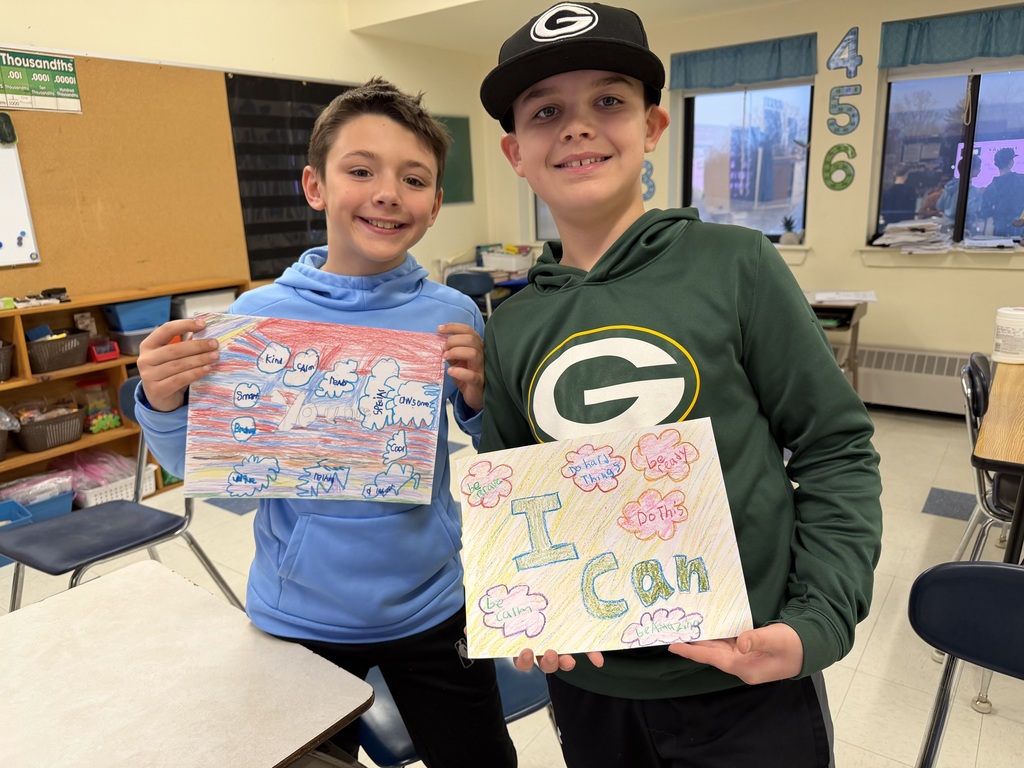 Two elementary students in a classroom smile while holding colorful hand-drawn posters with encouraging words and phrases like “I can.”