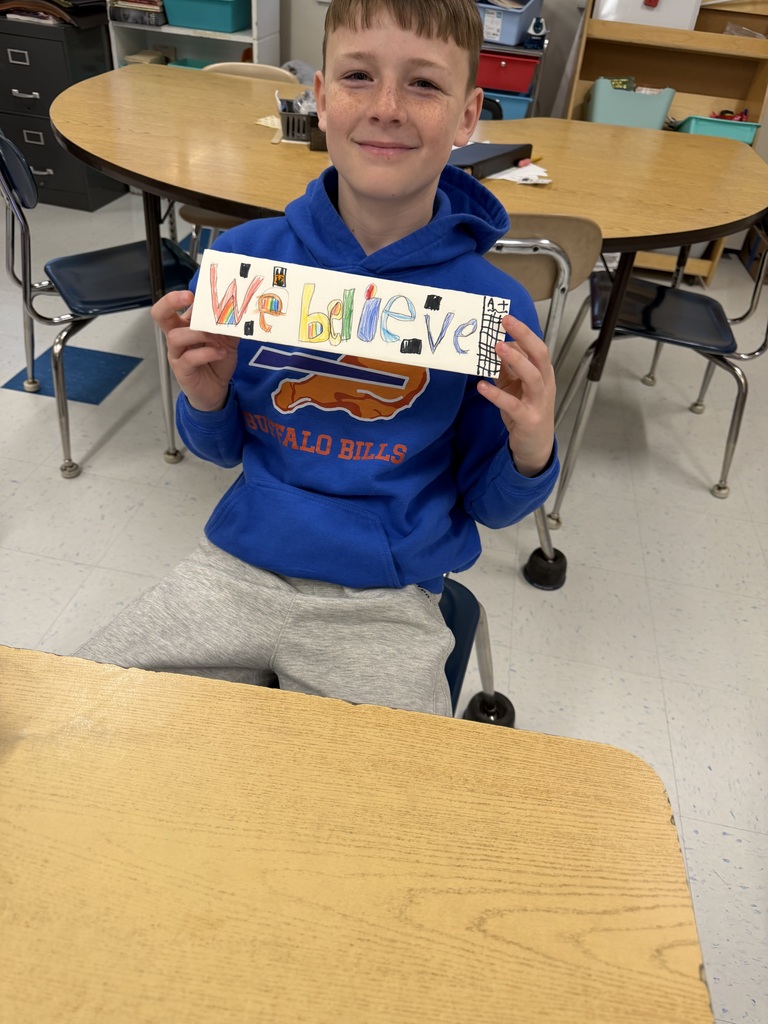 A student sits at a desk holding a handmade sign that reads “We believe,” decorated with colorful letters and drawings.