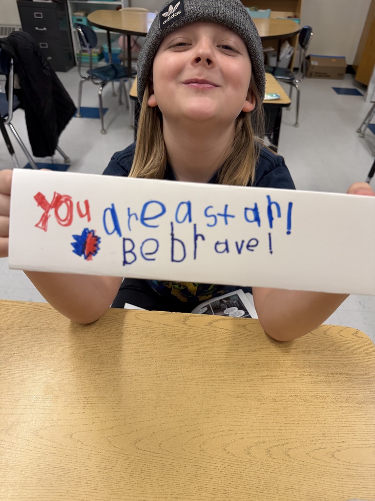 A student wearing a hat holds a sign that reads “You are a star! Be brave!” written in marker with simple drawings.