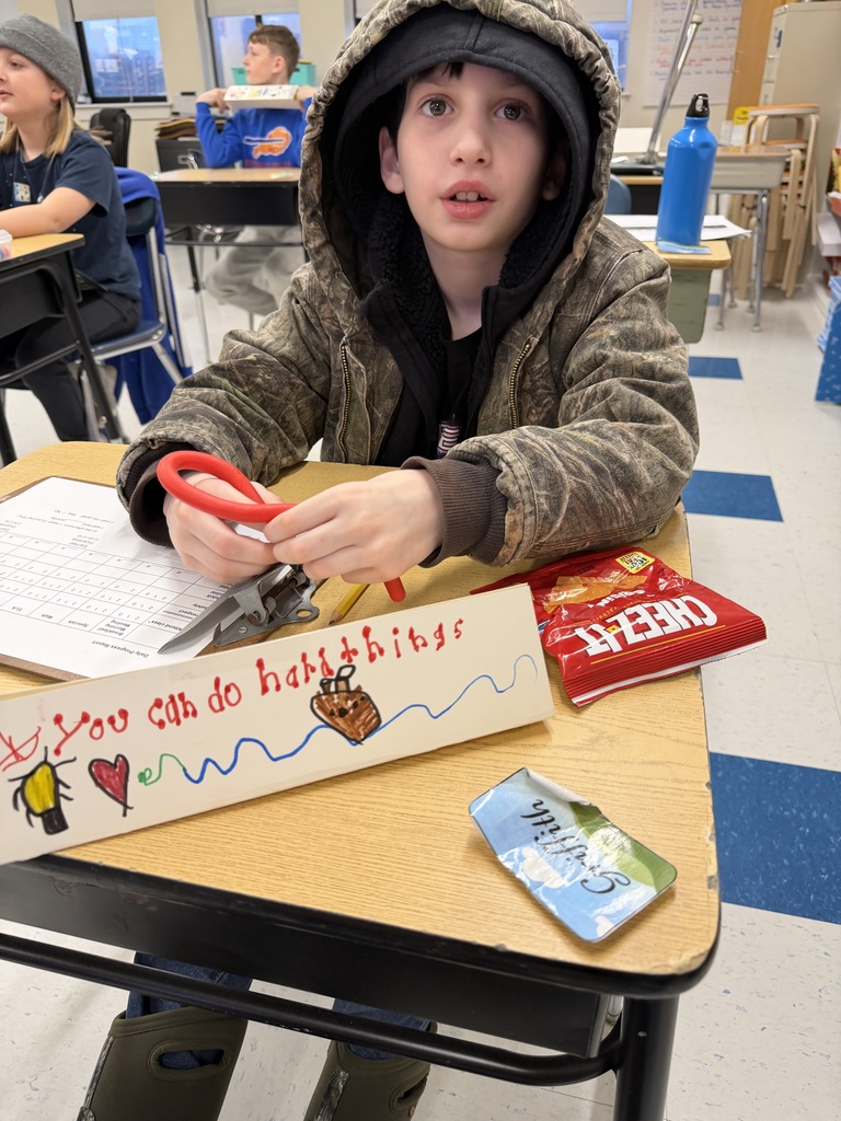 A student sits at a desk holding a handmade sign that reads “You can do hard things,” with drawings of a heart and other small illustrations nearby.
