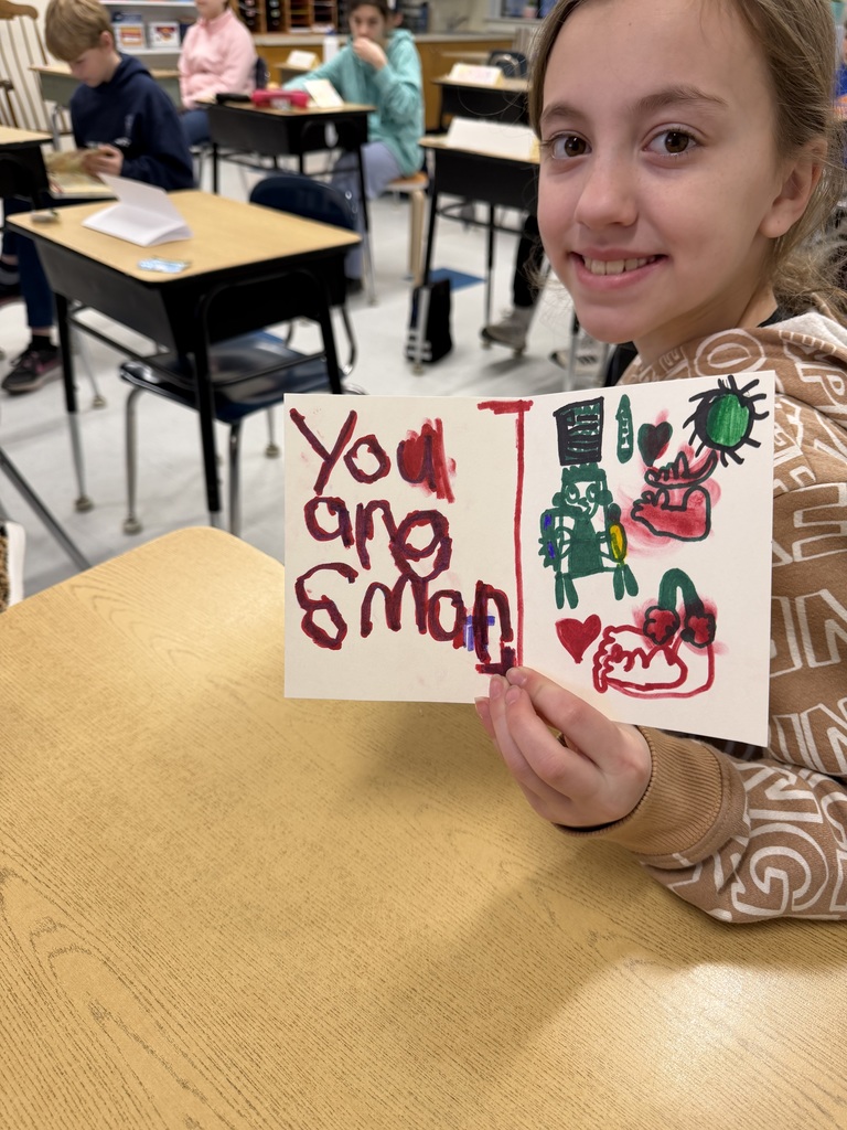 A student smiles while holding a card that says “You are smart,” decorated with drawings including hearts and figures.